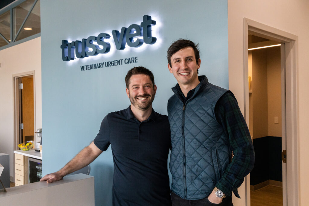 Brad Waffa (left) and CJ Casselli, co-founders of Truss Vet in Cary, North Carolina, stand beside a backlit dark blue sign of the company name in the lobby. Waffa wears a black polo shirt and Casselli a dark blue puffed jacket with a black long sleeve shirt underneath.